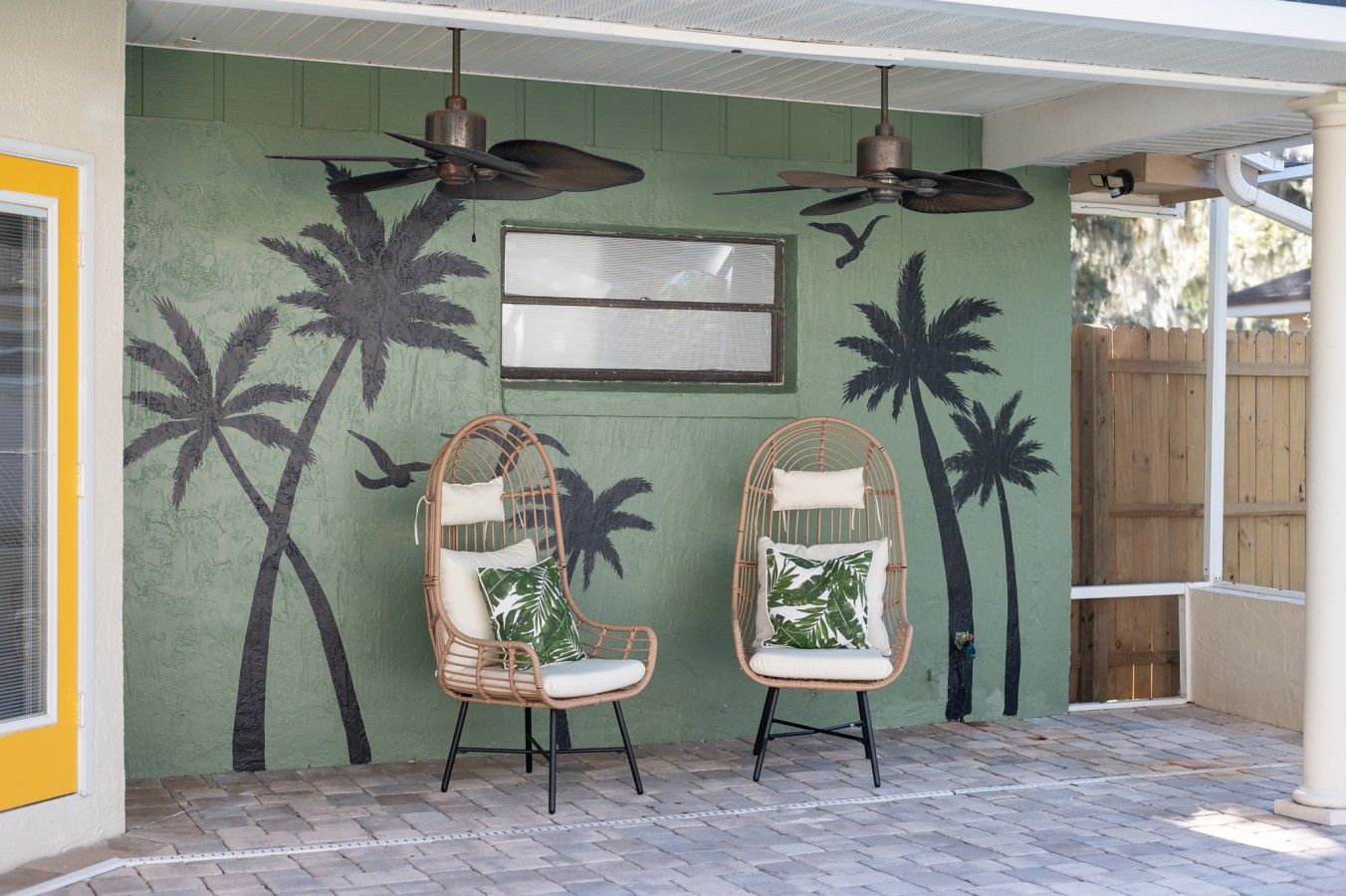 Un patio cubierto con dos sillas de mimbre, un ventilador de techo y un mural en la pared con palmeras.