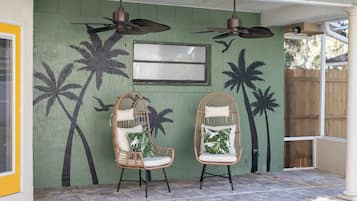 Un patio cubierto con dos sillas de mimbre, un ventilador de techo y un mural en la pared con palmeras.
