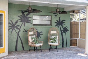 Un patio couvert comprenant deux fauteuils en osier, un ventilateur de plafond et une fresque murale représentant des palmiers.