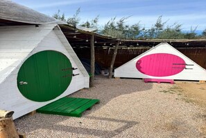 Dos pequeñas estructuras triangulares de madera con puertas circulares verdes y rosas, sobre una superficie de grava con una valla de madera y árboles al fondo.