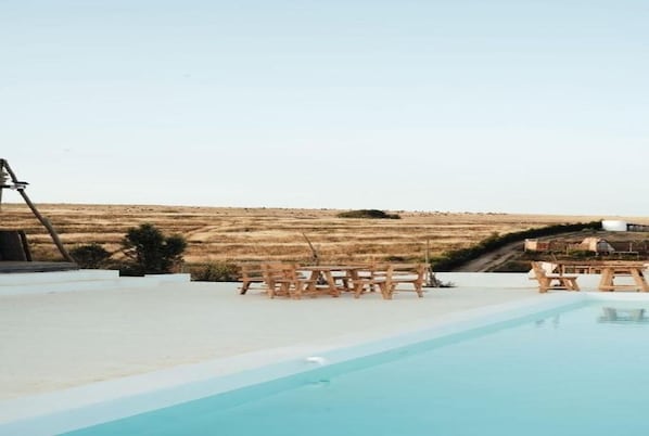 An outdoor swimming pool with wooden chairs and a table, surrounded by a white deck, with a view of a dry, hilly landscape.