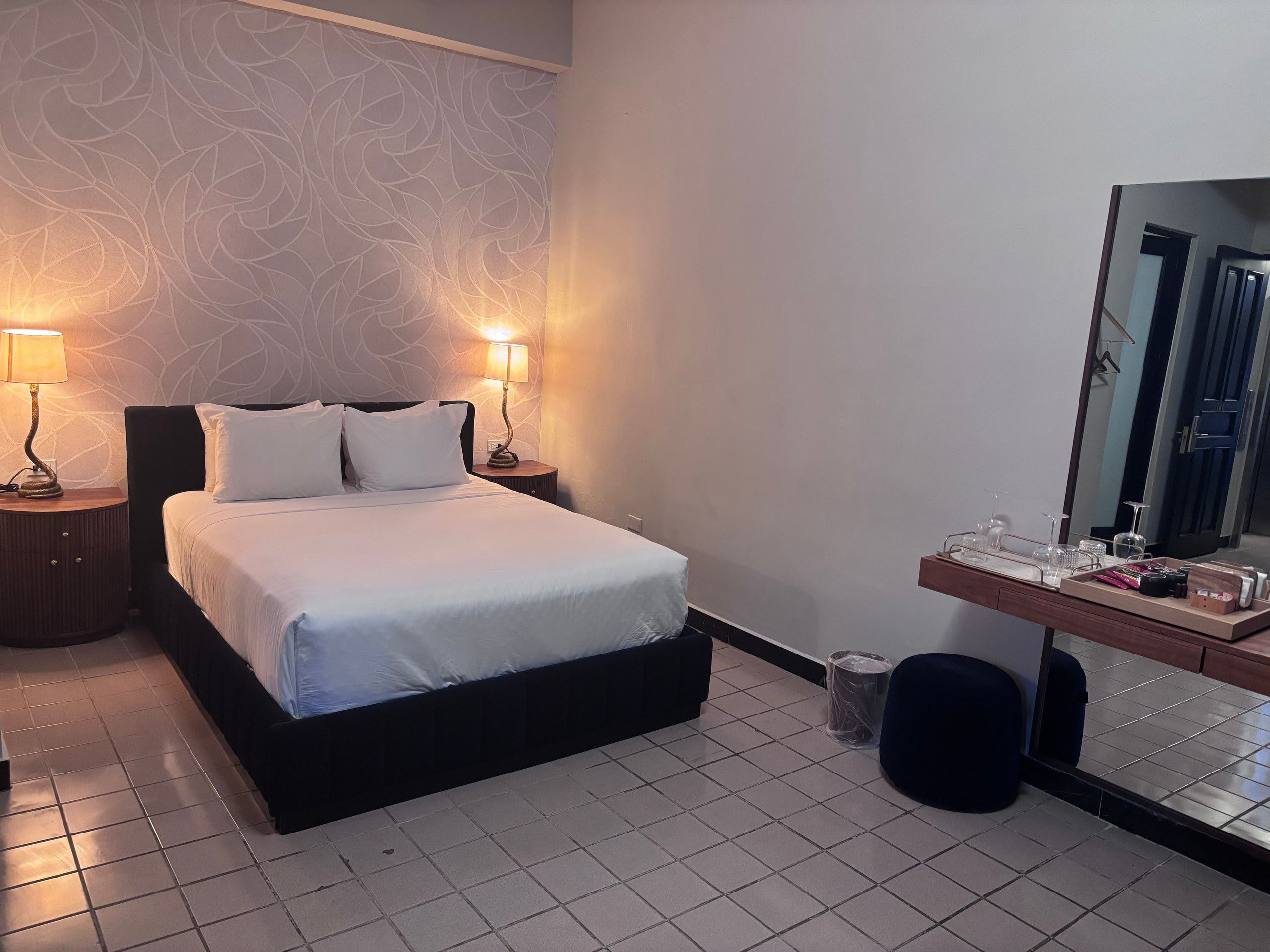 A hotel room with a bed, two bedside lamps, a dresser with toiletries, and a mirror.