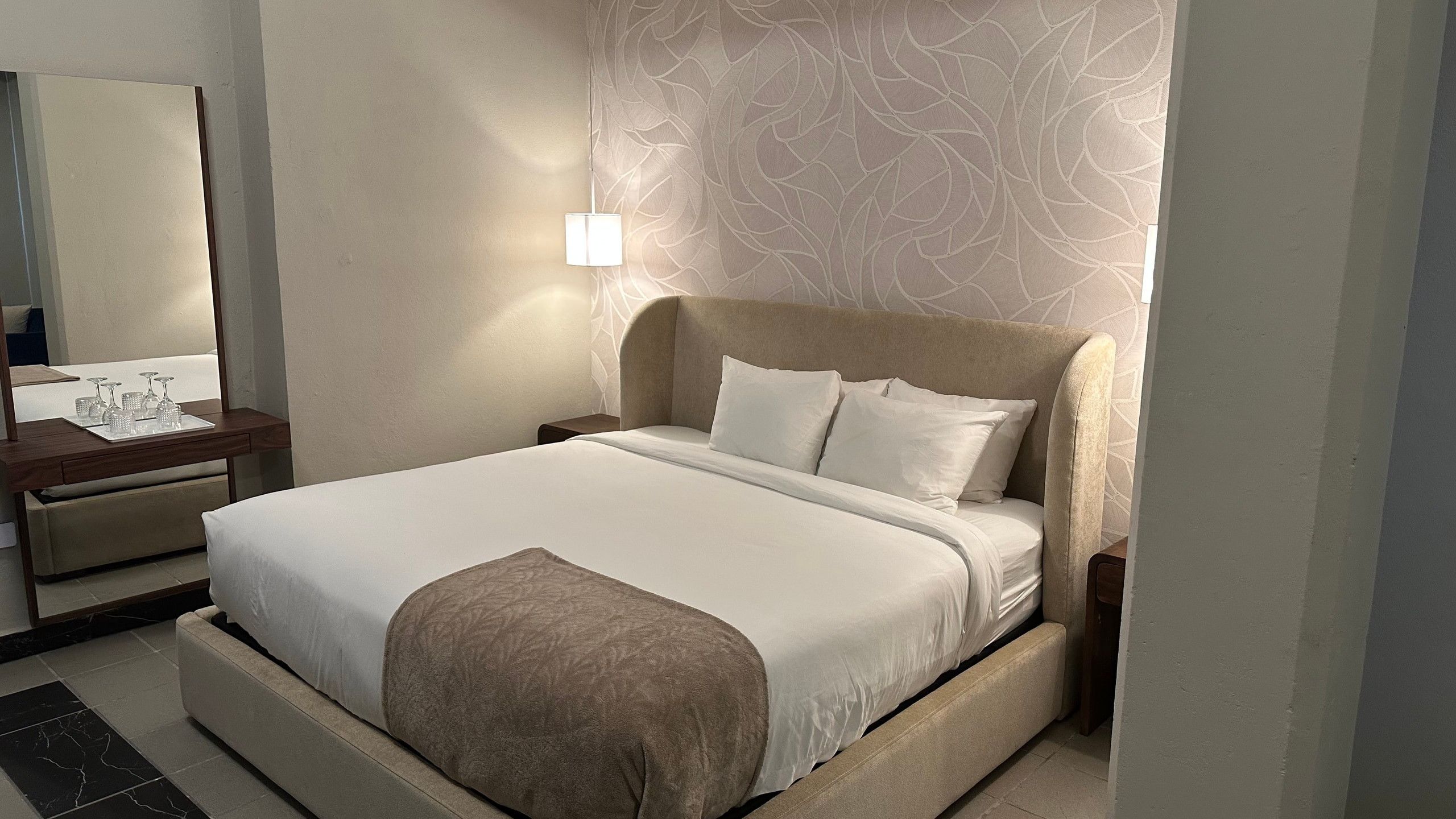A hotel room with a bed, bedside tables, a mirror, and a wall with a leaf pattern.