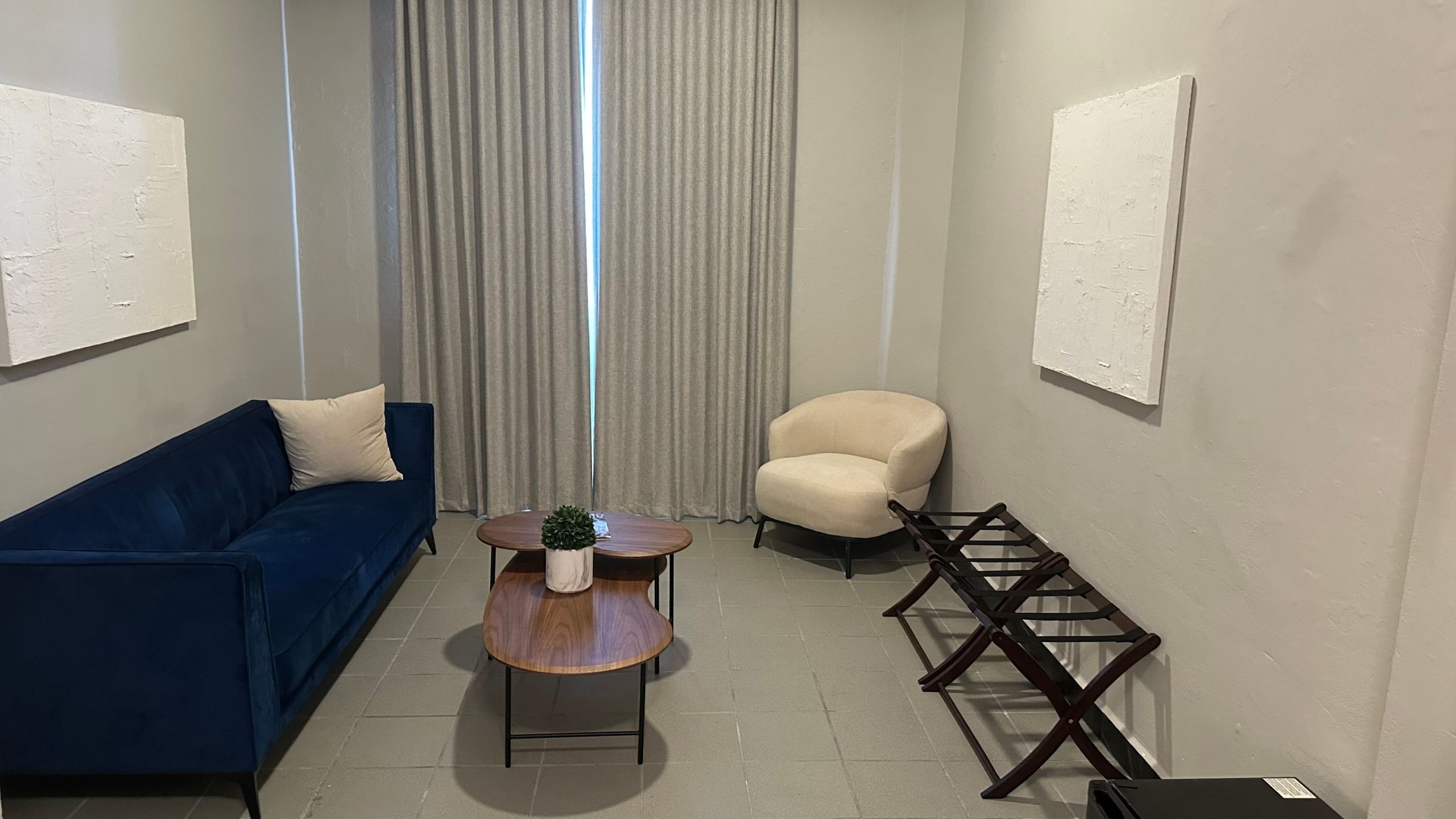 A room with a blue sofa, a round wooden coffee table with a plant, a beige armchair, and a wall-mounted rack.