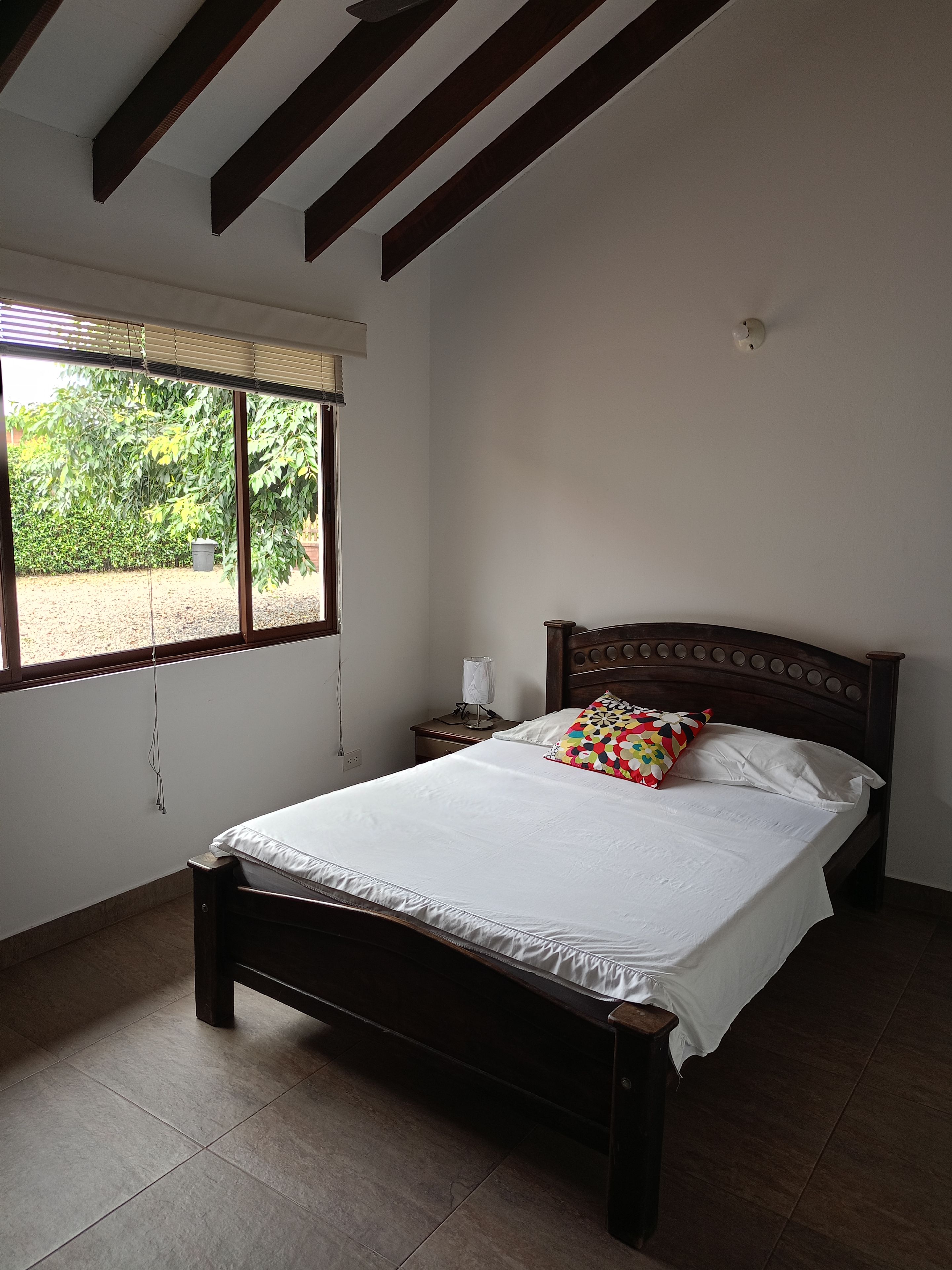 A bedroom with a wooden bed, white bedding, a nightstand with a lamp, and a window with blinds.