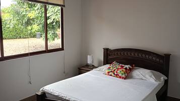 A bedroom with a wooden bed, white bedding, a nightstand with a lamp, and a window with blinds.