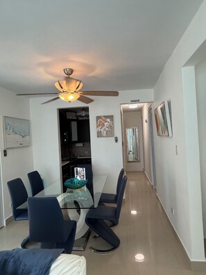 A modern dining area with a glass table, dark chairs, a ceiling fan, and a kitchen in the background. - New Listing (San Juan)