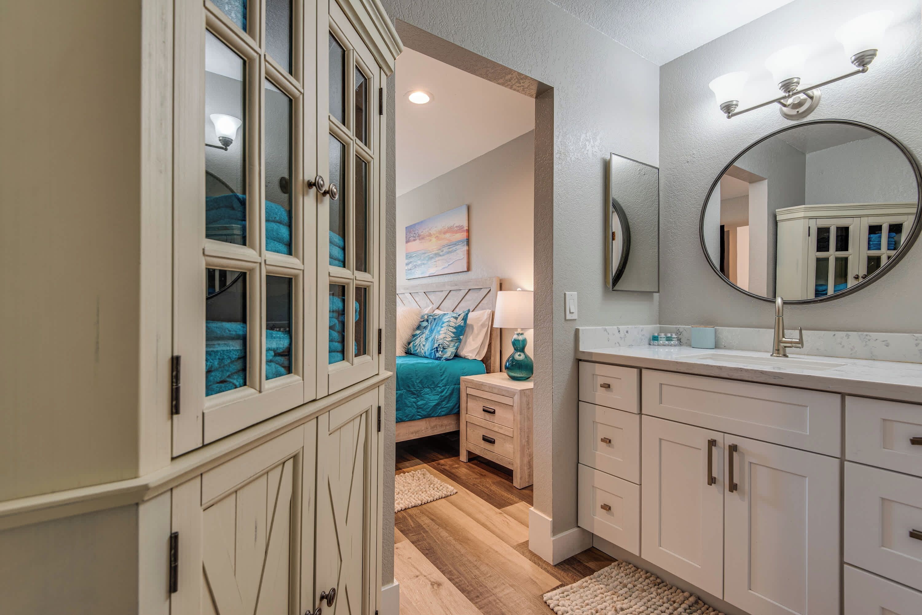 A bedroom with a large window, a bed with blue bedding, a nightstand, a mirror, and a vanity with a sink.