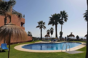 A swimming pool with a surrounding grass area, palm trees, and a building with a thatched roof. - Penthouse 'Doña Julia' with Sea View and Golf View (Casares)
