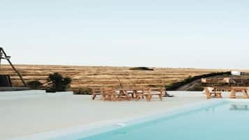 Piscina exterior con sillas y una mesa de madera, rodeada por una terraza blanca, con vistas a un paisaje seco y montañoso.