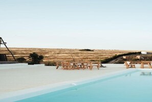 Piscina exterior con sillas y una mesa de madera, rodeada por una terraza blanca, con vistas a un paisaje seco y montañoso.