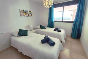 Quarto de hotel com duas camas, uma mesinha de cabeceira, luminária, pintura e janela com cortinas.