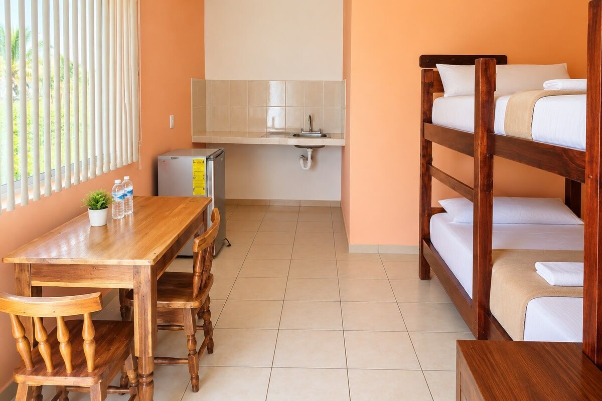 A dormitory room with bunk beds, a wooden desk, chairs, a small kitchenette, and a window with blinds.