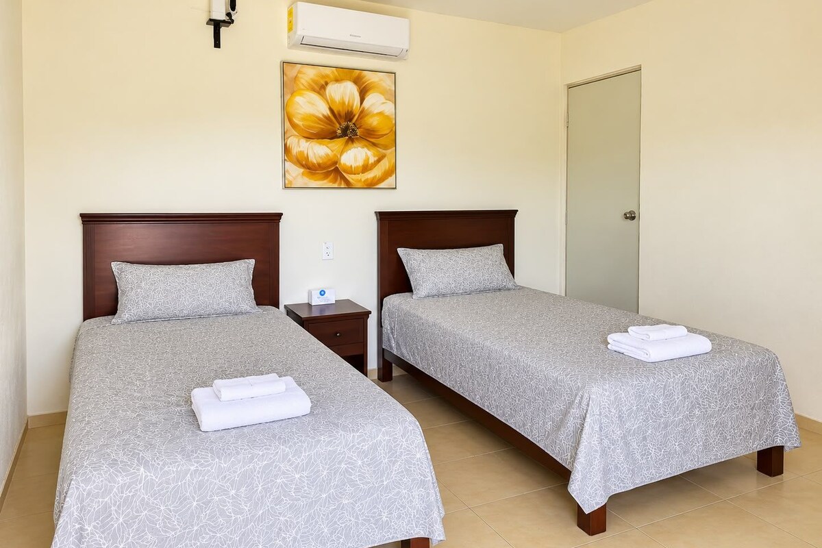 A hotel room with two beds, a wall-mounted air conditioner, a painting of a flower, and a door.