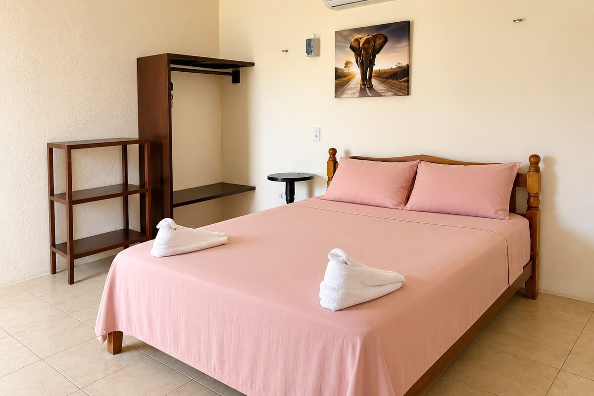 A hotel room with a bed, a wooden wardrobe, a small table, and an air conditioning unit.