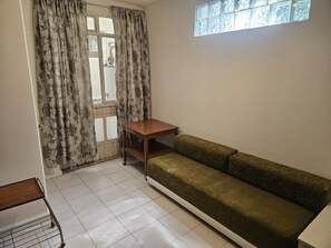 Ein Zimmer mit einer Couch, einem kleinen Tisch, einem Fenster mit gemusterten Vorhängen und einem gefliesten Boden.