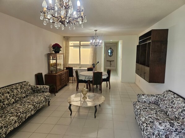 A living room with a chandelier, floral sofas, a dining table with chairs, and a cabinet with glass doors.