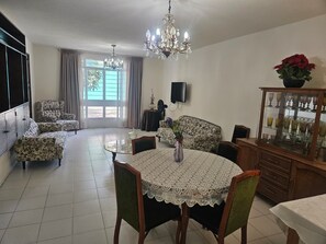 A living room with a dining table, chairs, a sofa, a chandelier, and a cabinet with glassware.