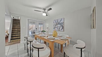 Uma área de refeições com uma mesa de madeira posta para seis, cadeiras brancas, um ventilador de teto e um quadro na parede.