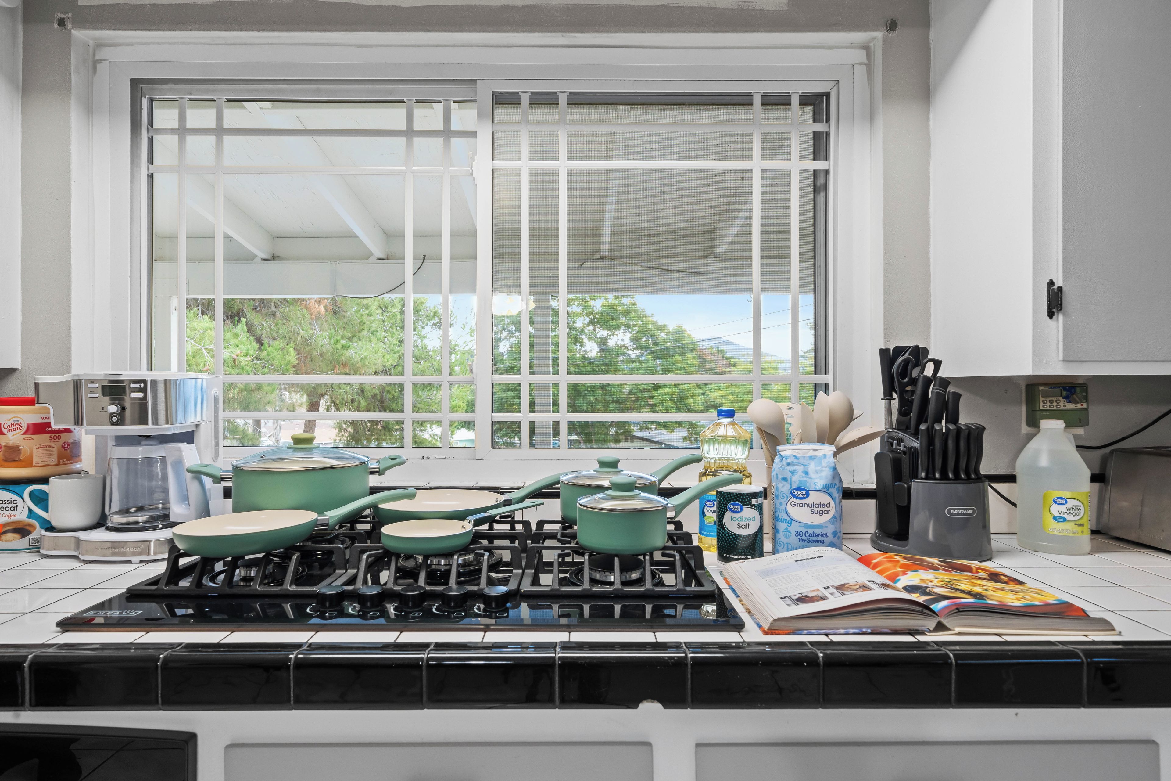 Cozinha com fogão a gás, panelas, cafeteira e janela com vista para árvores.