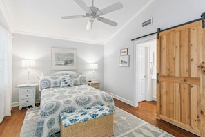 Une chambre à coucher comprenant un lit, des tables de chevet, un ventilateur de plafond, une porte en bois et un mur orné de tableaux encadrés.