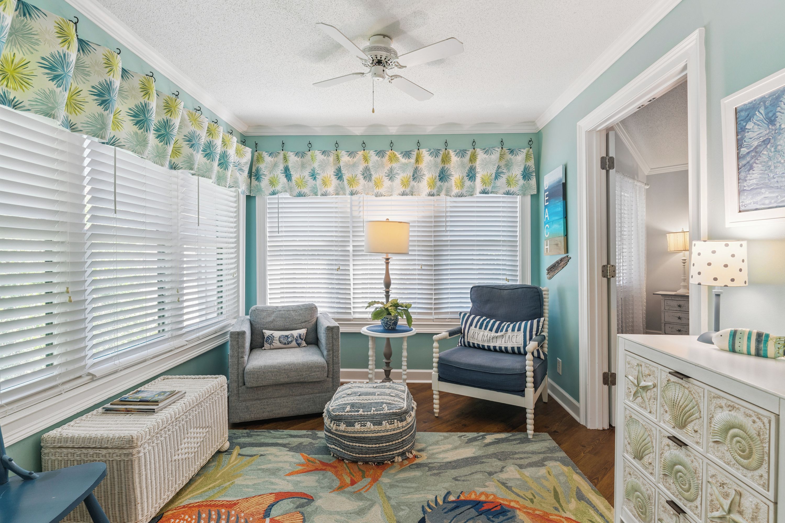 A room with a ceiling fan, two windows with white blinds, a grey armchair, a blue armchair, a small round table with a lamp, a wicker ottoman, and a decorative rug with a fish design.