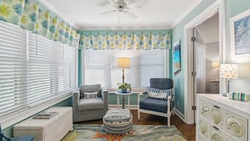 A room with a ceiling fan, two windows with white blinds, a grey armchair, a blue armchair, a small round table with a lamp, a wicker ottoman, and a decorative rug with a fish design.
