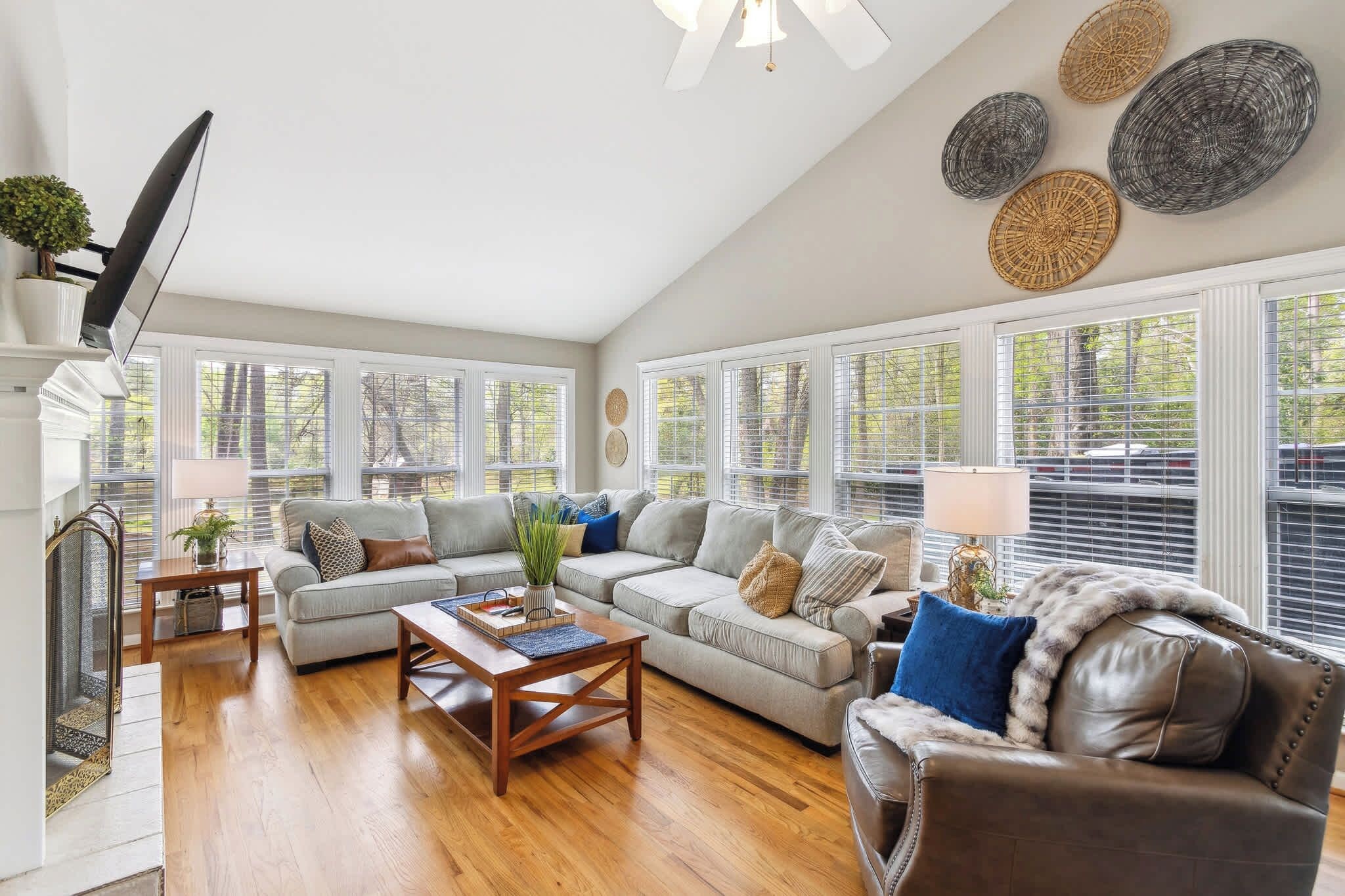 A spacious living room with a large sectional sofa, a leather armchair, and a wooden coffee table. There are large windows with blinds, a fireplace, and decorative wall art.