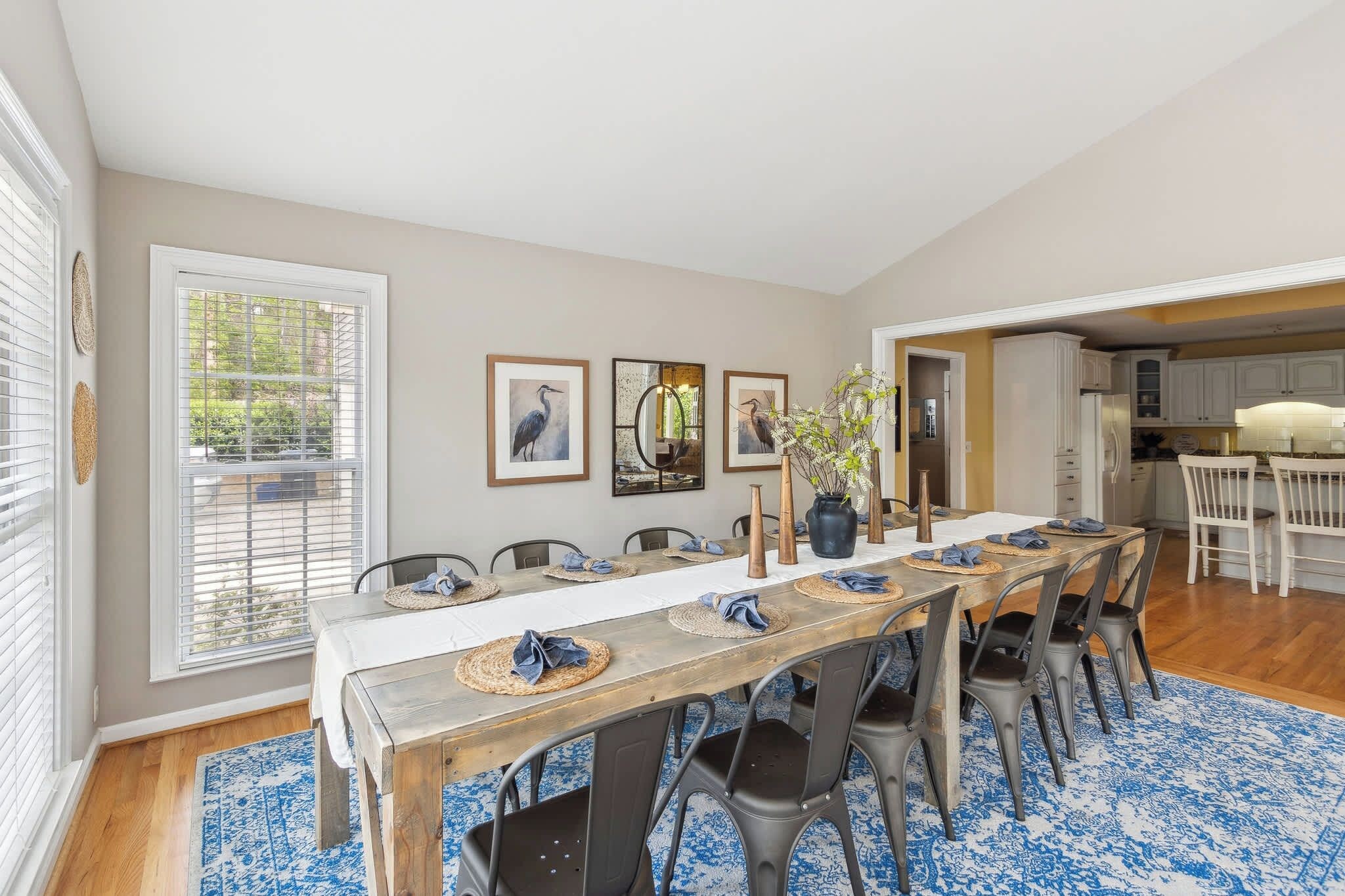 A dining area with a long table set for six, flanked by chairs, with a kitchen visible in the background.