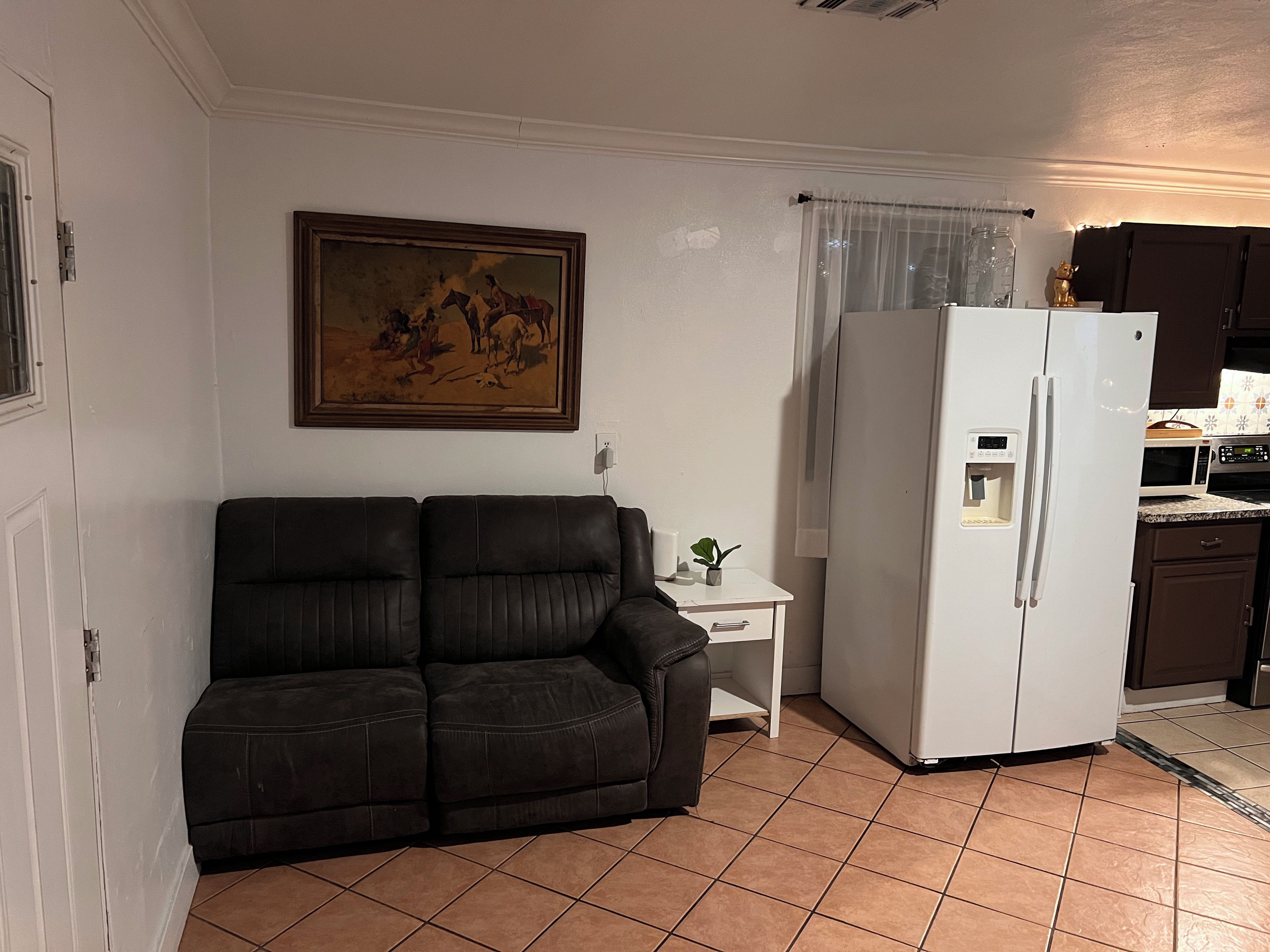 Ett vardagsrum med en mörkbrun soffa, ett vitt sidobord med en växt och ett vitt kylskåp.