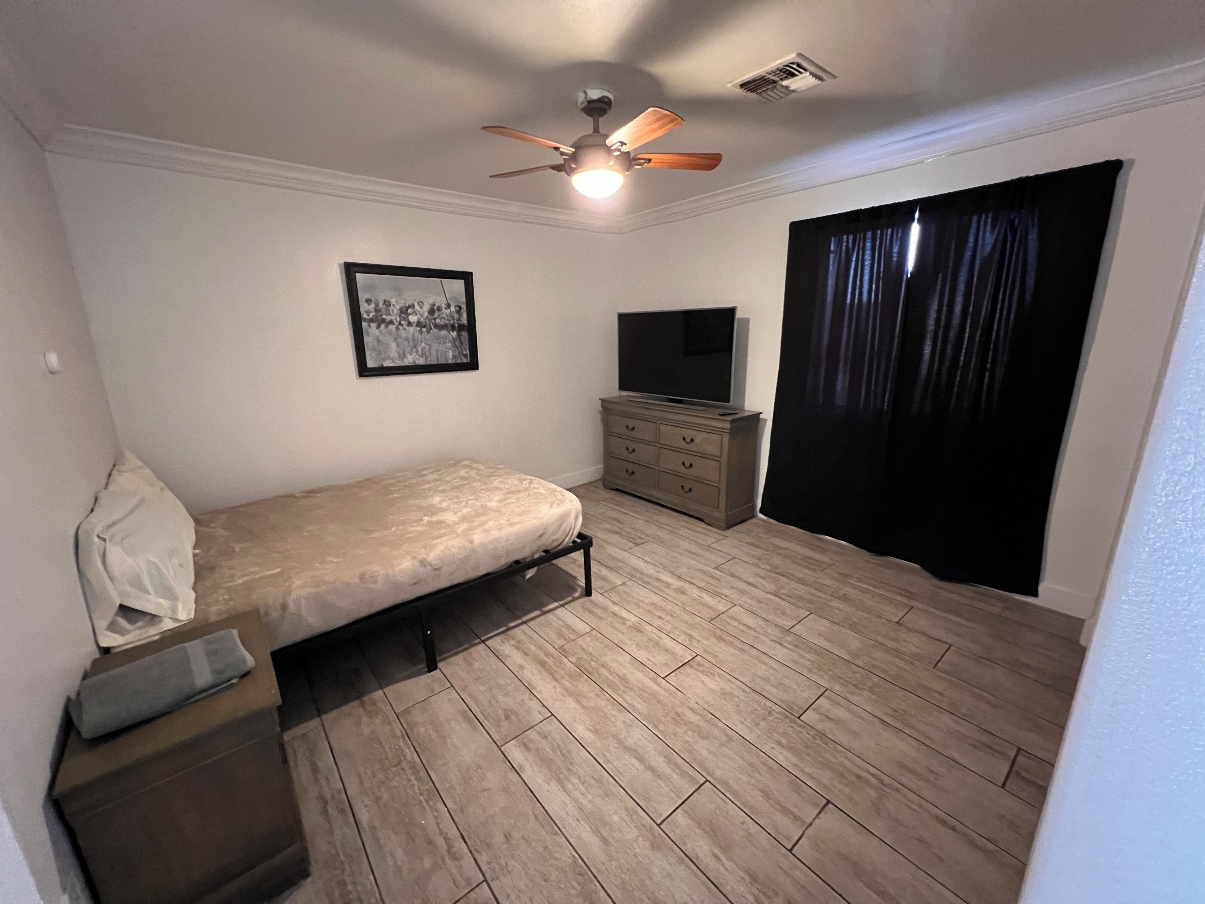 A bedroom with a bed, a dresser, a television, a ceiling fan, and a window with curtains.