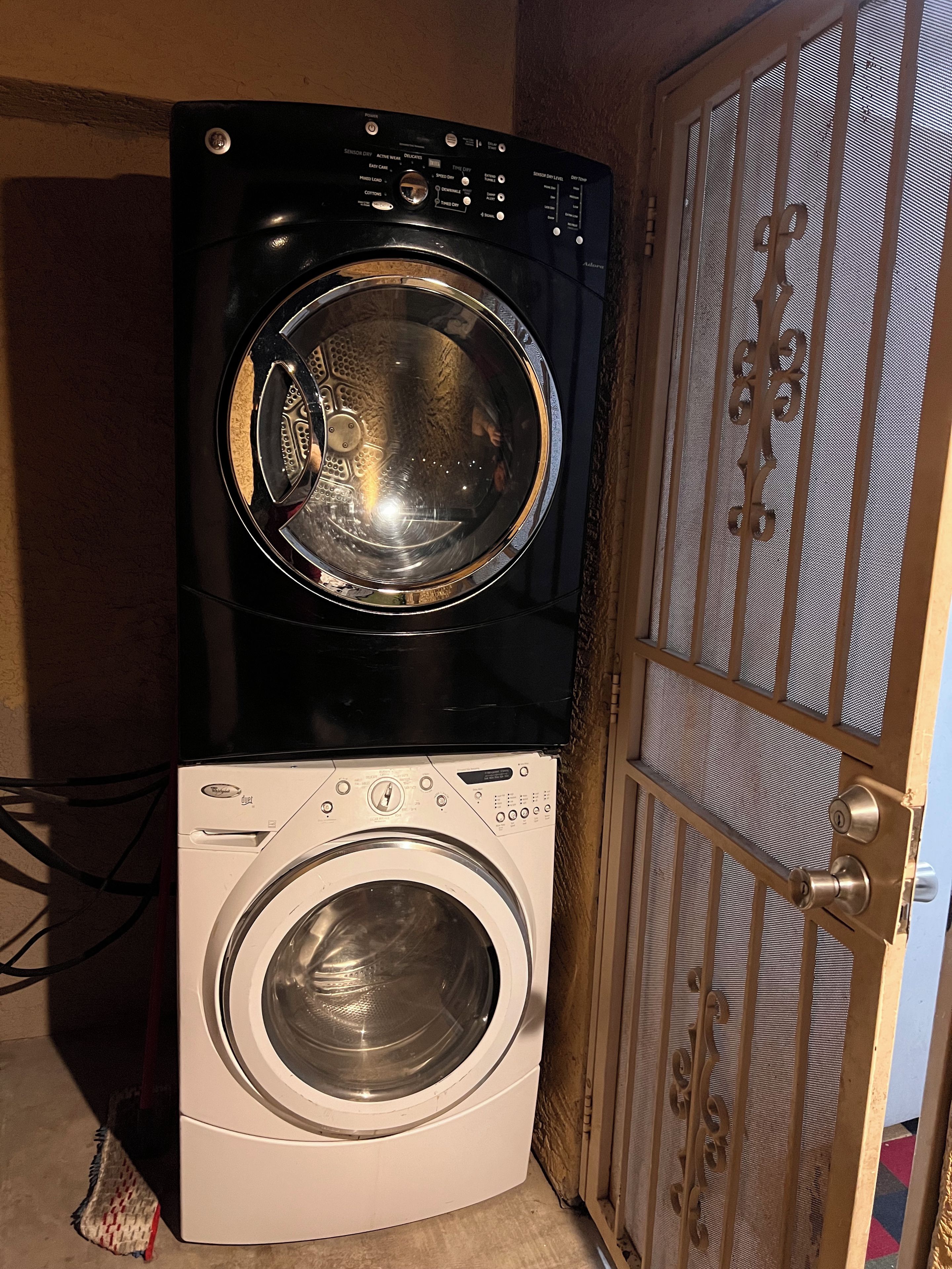 A front-loading washing machine and dryer stacked in a laundry room with a wooden door and metal mesh screen.