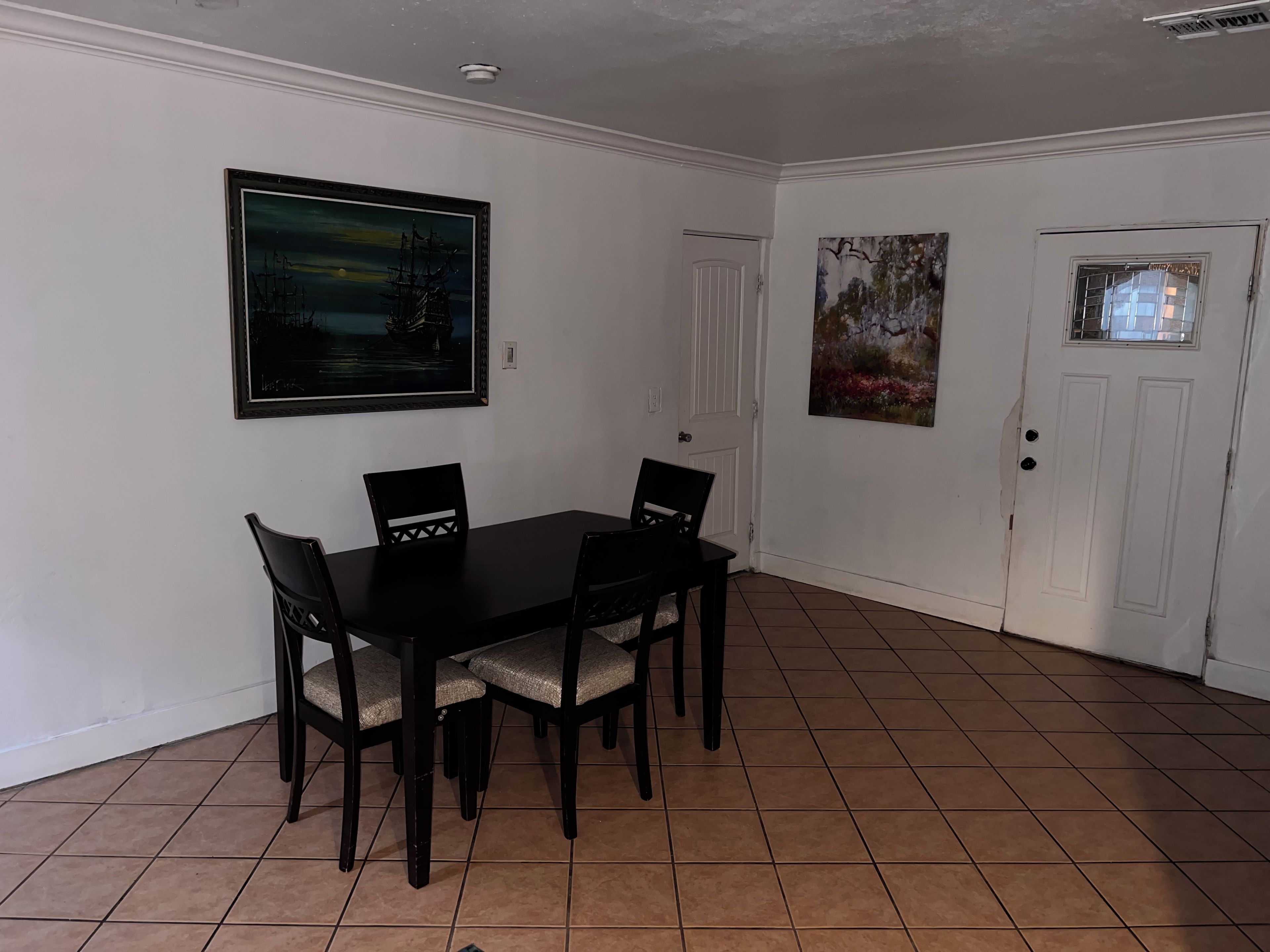 Un coin repas comprenant une table rectangulaire et quatre chaises, une porte blanche et deux tableaux accrochés au mur.