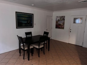 Una zona de menjador amb una taula rectangular i quatre cadires, una porta blanca i dos quadres penjats a la paret.