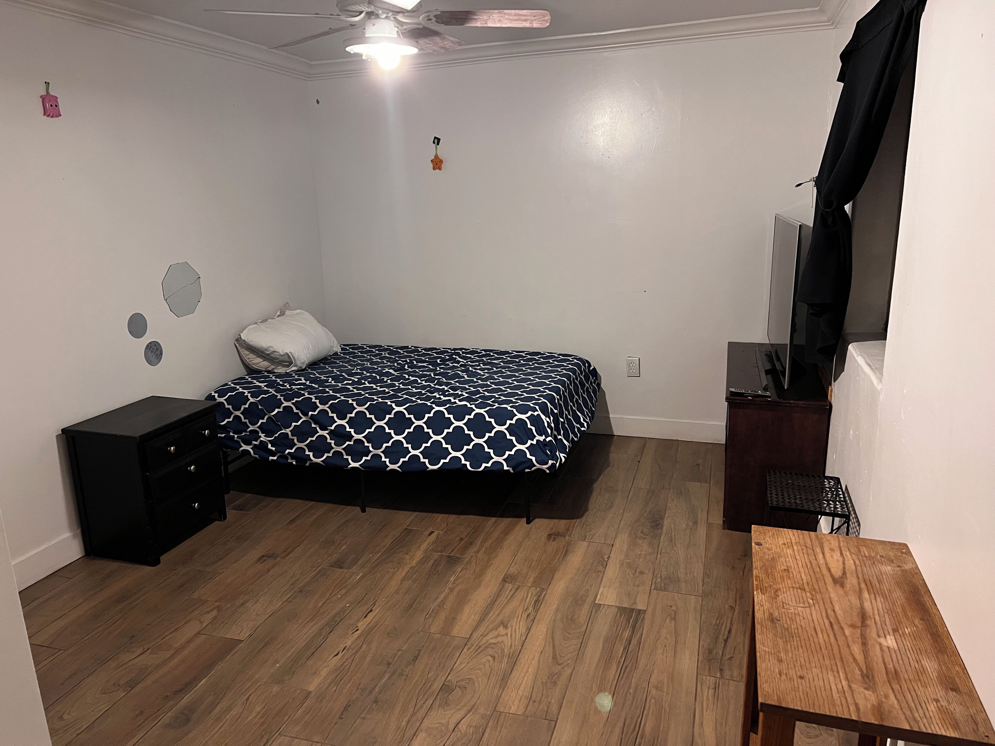 A bedroom with a bed, a nightstand, a wooden table, a ceiling fan, and a wall-mounted TV.
