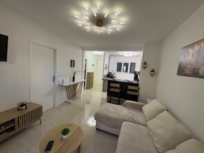 Ett modernt vardagsrum med en soffa, ett soffbord och en tv. Det finns en matplats med en bardisk och stolar. Rummet har en taklampa och en dörr.