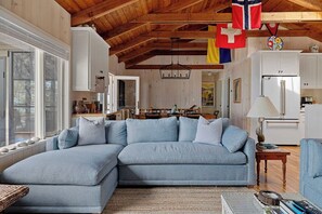 A living room with a sectional sofa, wooden ceiling beams, white cabinets, and a dining area in the background. - The Lake House | Private Beach • Sunsets • 6BR! (Bridgman)