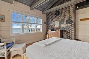 A bedroom with a large window, a bed, a dresser, and a brick wall. - The Lake House | Private Beach • Sunsets • 6BR! (Bridgman)