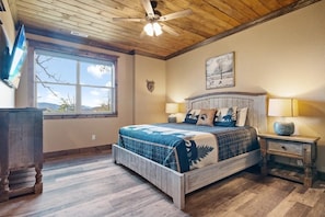 A bedroom with a wooden ceiling, a bed with a blue and white patterned bedspread, a headboard, two bedside lamps, and a window with a view of trees and mountains. - Star of the Summit: 5 bedrooms, Private Indoor Pool, Stunning Views (Sevierville)