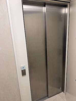 Suljettu hissi, jonka pinnassa on metallinhohtoa, seinässä oleva ohjauspaneeli ja laatoitettu lattia.