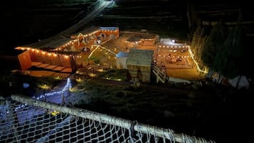 Un campamento con tiendas de campaña y una estructura de madera, rodeado por una valla y iluminado con luces colgantes.