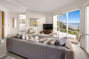 A modern living room with a large sofa, a television, and a view of the sea.