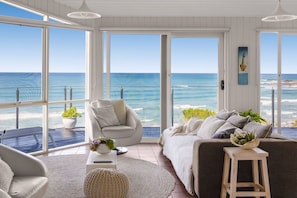 A room with a large sliding glass door leading to a balcony with a view of the ocean. There is a white sofa, a small round table, and a potted plant on the balcony.