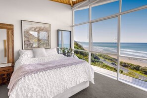 A bedroom with a large window overlooking a beach and ocean, a bed with white bedding, and a wooden headboard.