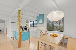 A modern kitchen with a central island, a dining area with a wooden table, and a large window with a view of trees. - Jan Juc beach retreat: gentle 5 min walk to beach, ocean views and fire pit. (Jan Juc)