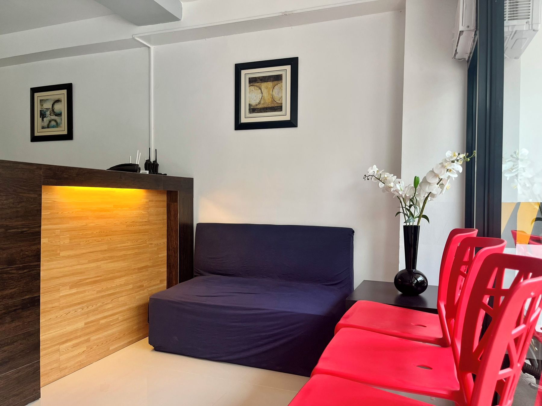 A modern room with a wooden reception desk, a blue sofa, red chairs, and a vase with white flowers.