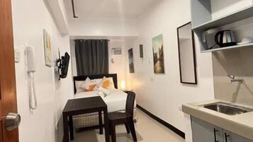 A compact hotel room with a bed, bedside table, and a small kitchenette.