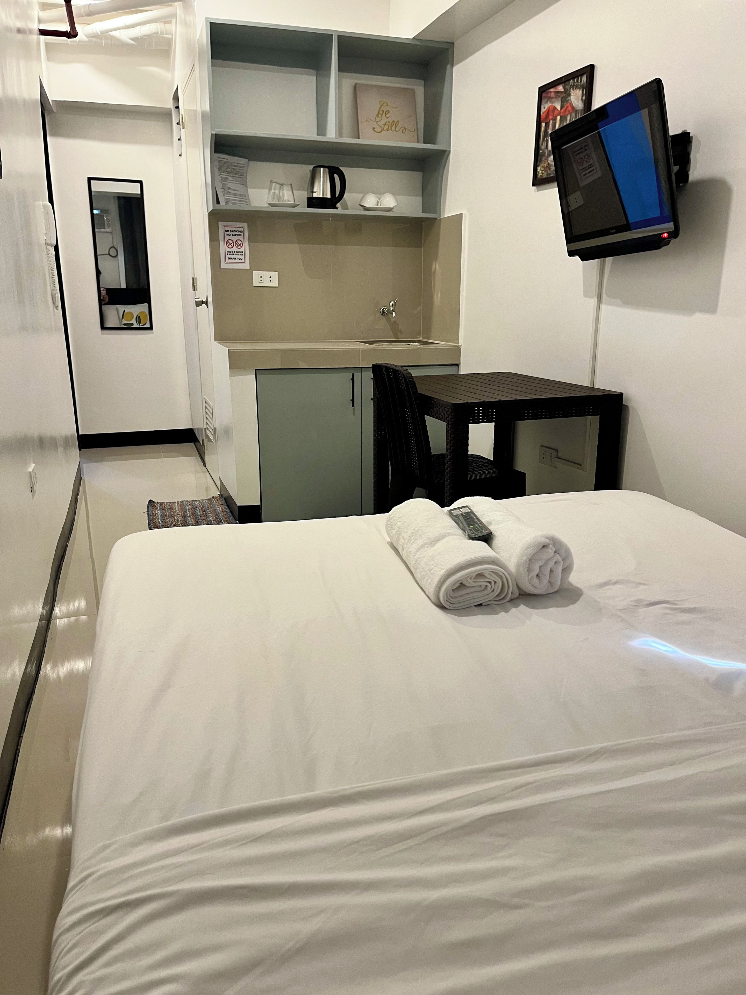 Habitación de hotel con cama, escritorio, kitchenette pequeña y televisor montado en la pared.