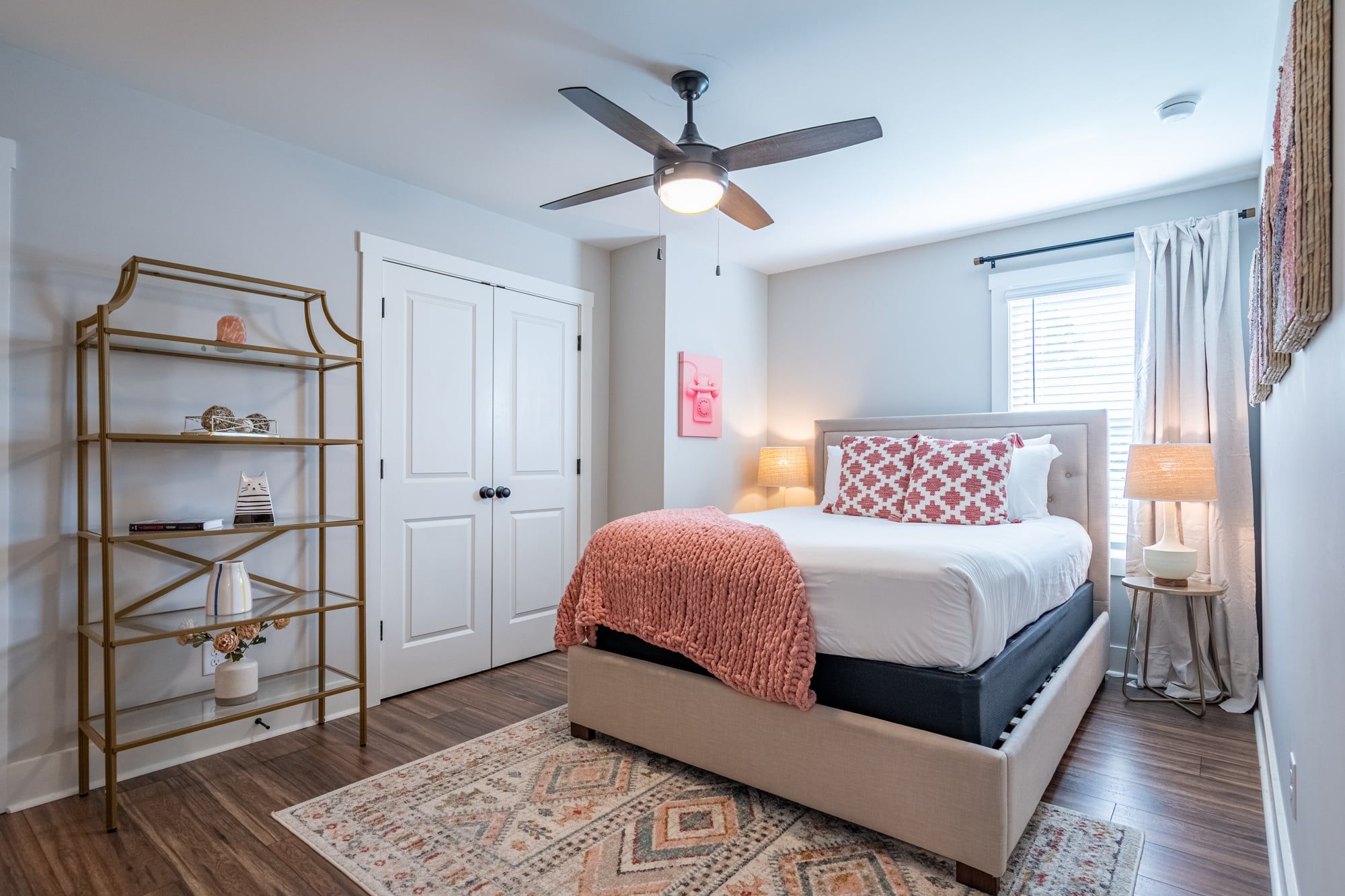 A bedroom with a bed, a ceiling fan, a white wardrobe, a shelf unit, and a patterned rug.