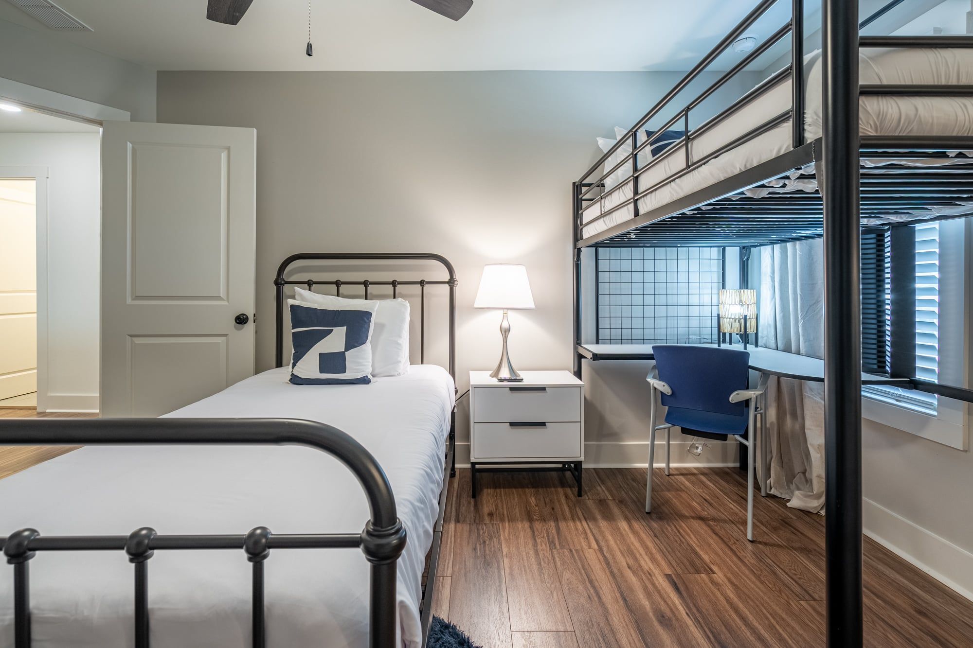 A bedroom with a bunk bed, a bedside table with a lamp, a chair, and a window with blinds.